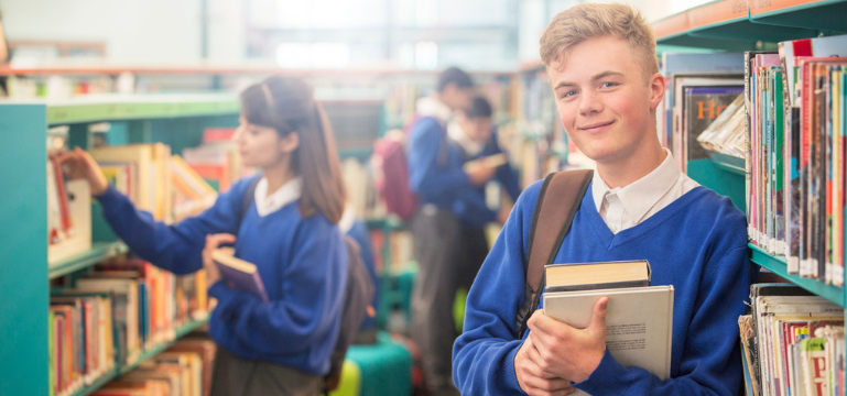 Two students in blue school uniforms browsing books in a library, one smiling at the camera while holding books.