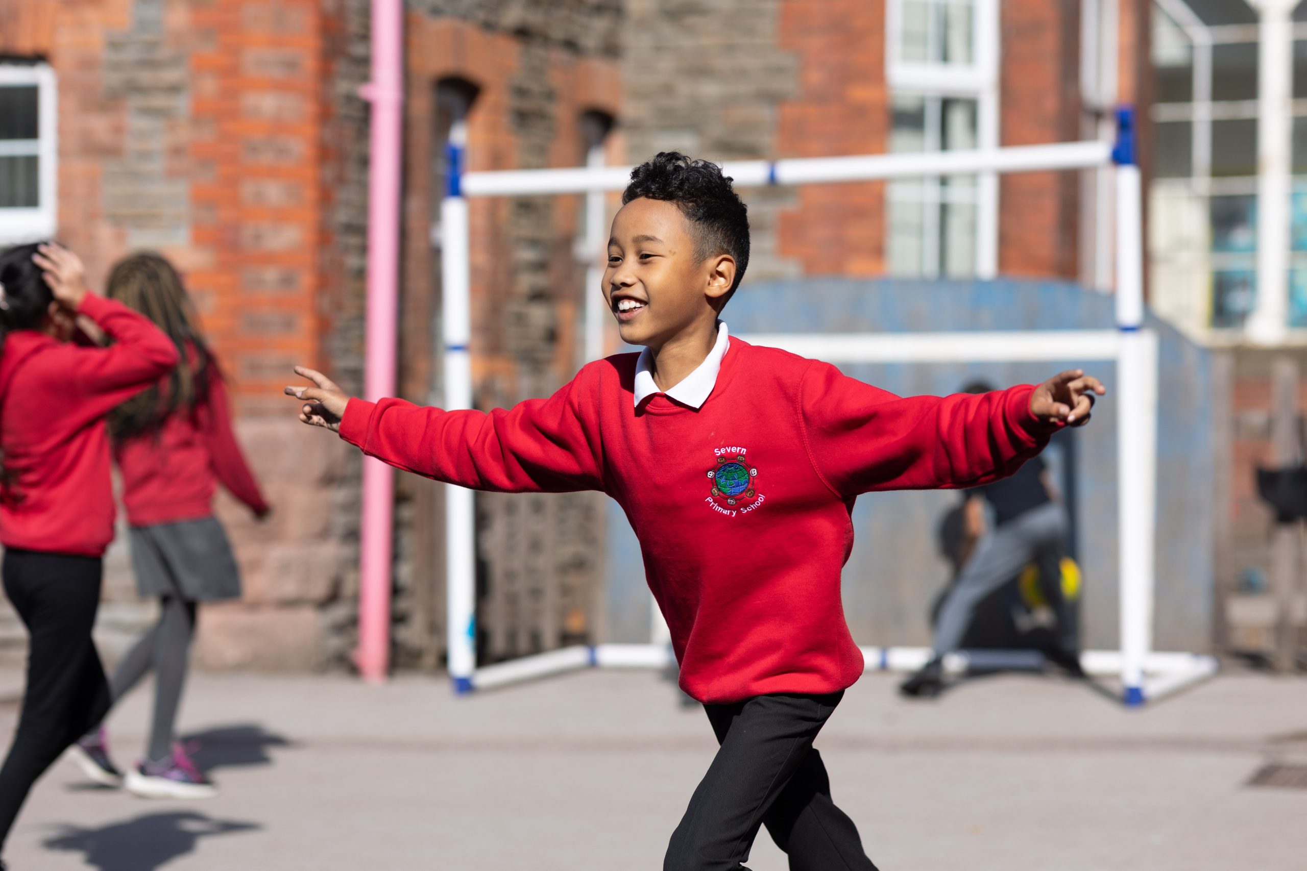 A young boy in a red school jumper is playing outside, arms outstretched and smiling, with a brick school building in the background.