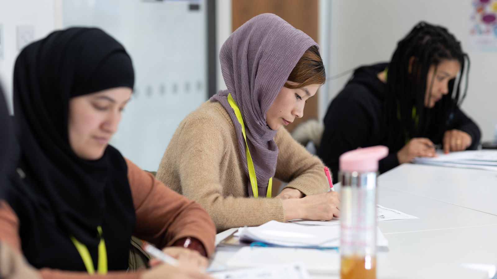 A woman writing in a notebook at a desk, with other people working in the background.