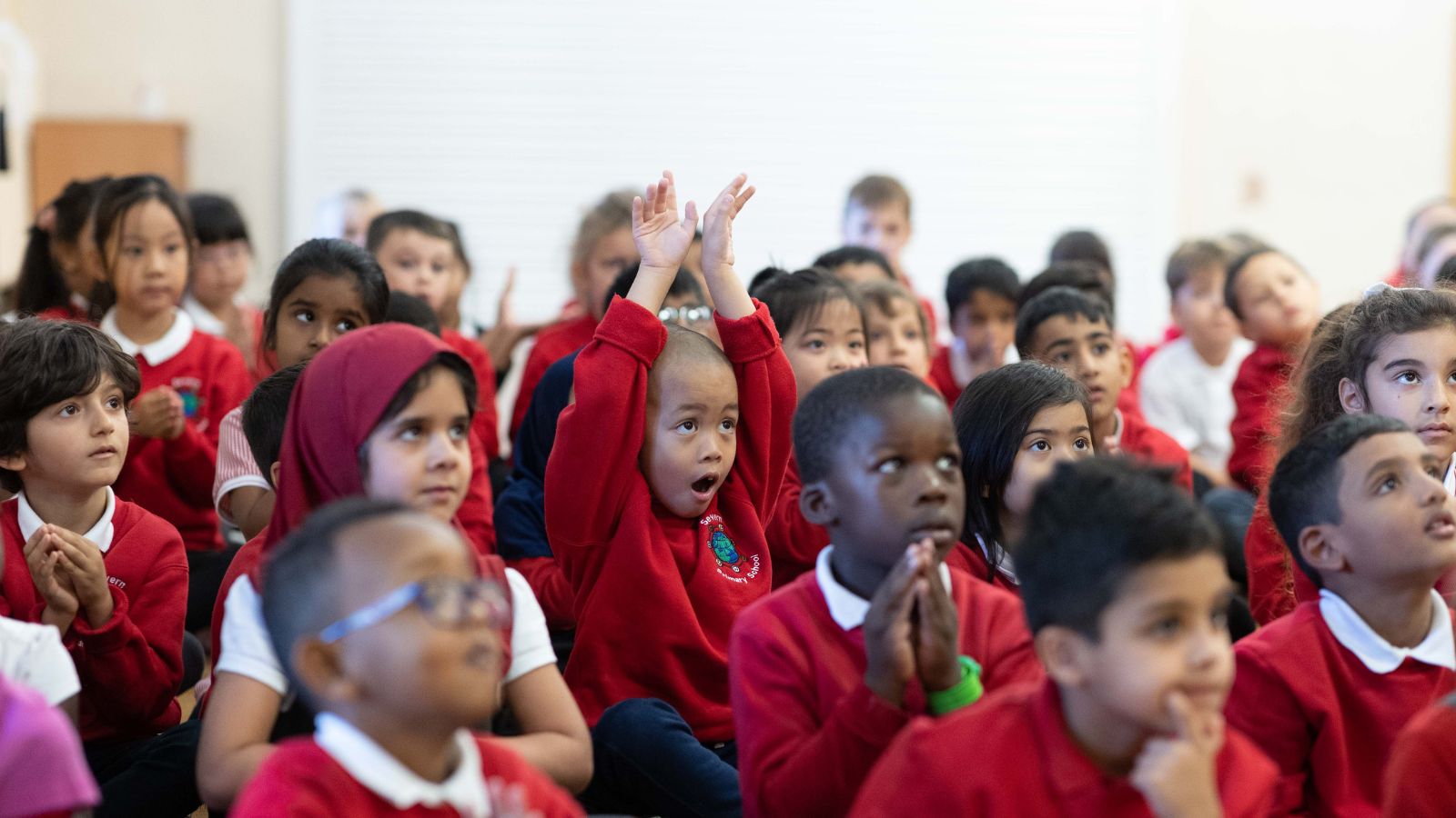 A group of young children sitting together, wearing red school uniforms, with one child raising their hands and looking excited.