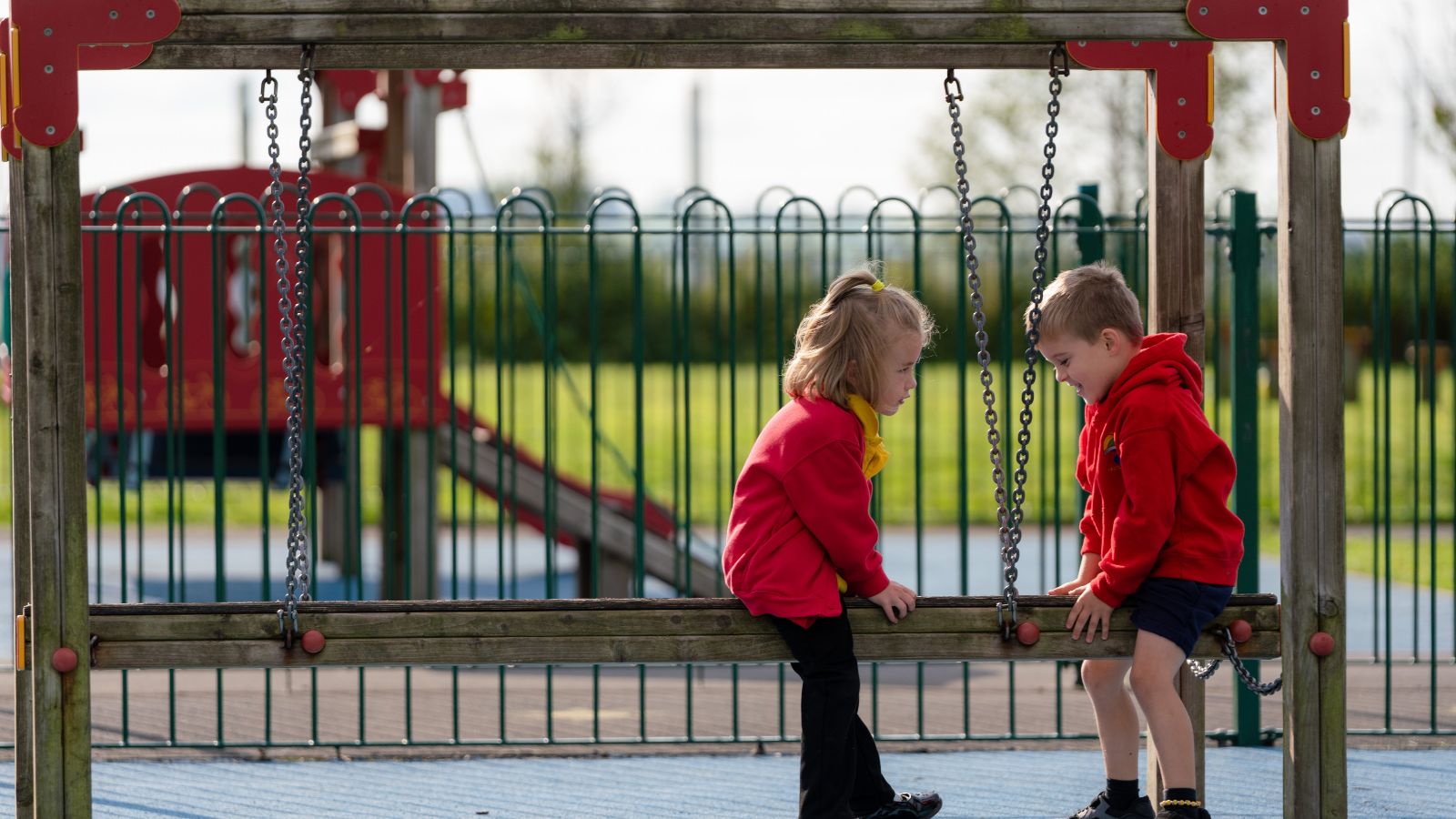 Two young children wearing red hoodies playing on a wooden climbing frame in a playground.