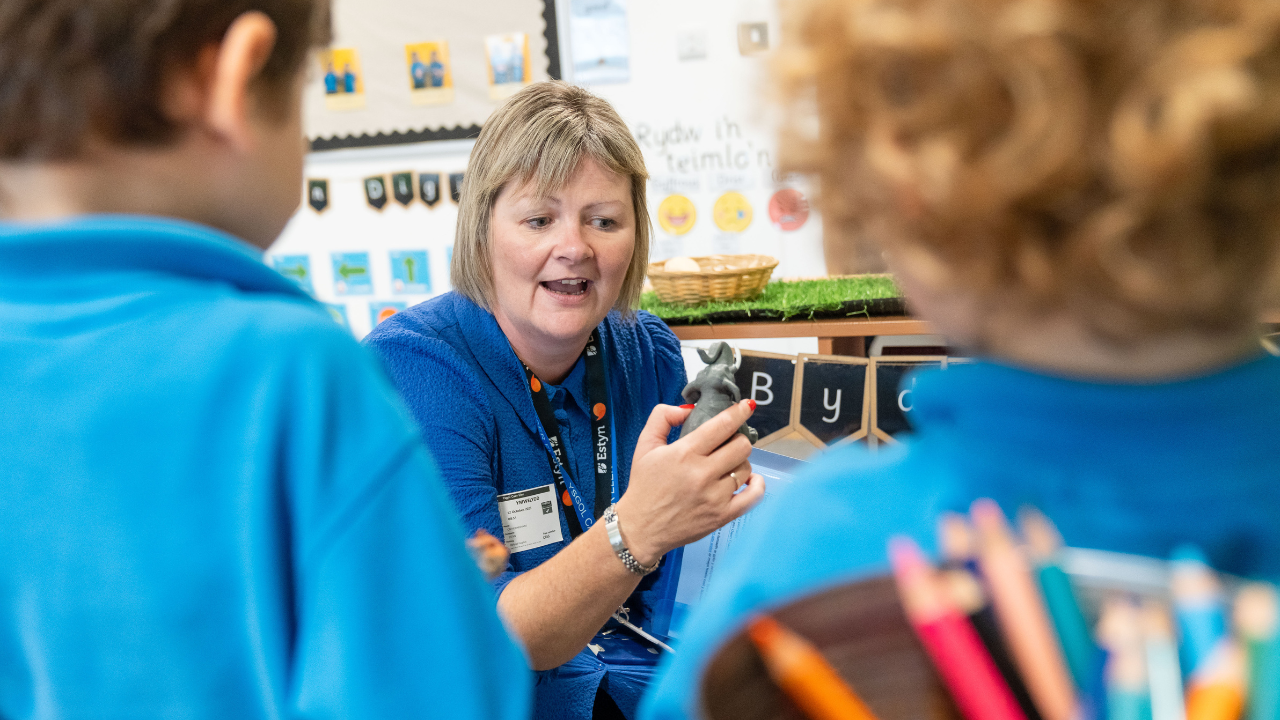 A woman in a blue shirt sitting on the floor, holding an object and talking to two children. The classroom is decorated with educational materials and children's artwork.