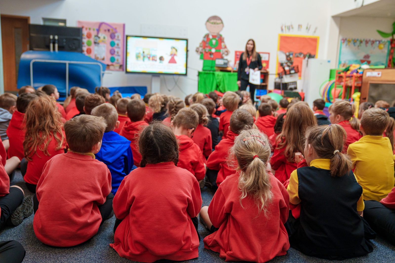 A large group of young children in red school uniforms sitting on the floor, facing a teacher who is giving a presentation in a colourful classroom.