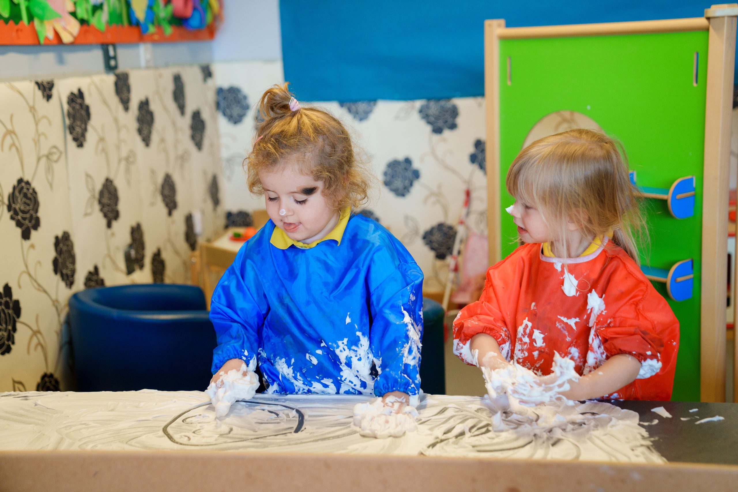 Two young children playing with shaving foam on a table in a classroom, wearing colorful aprons.