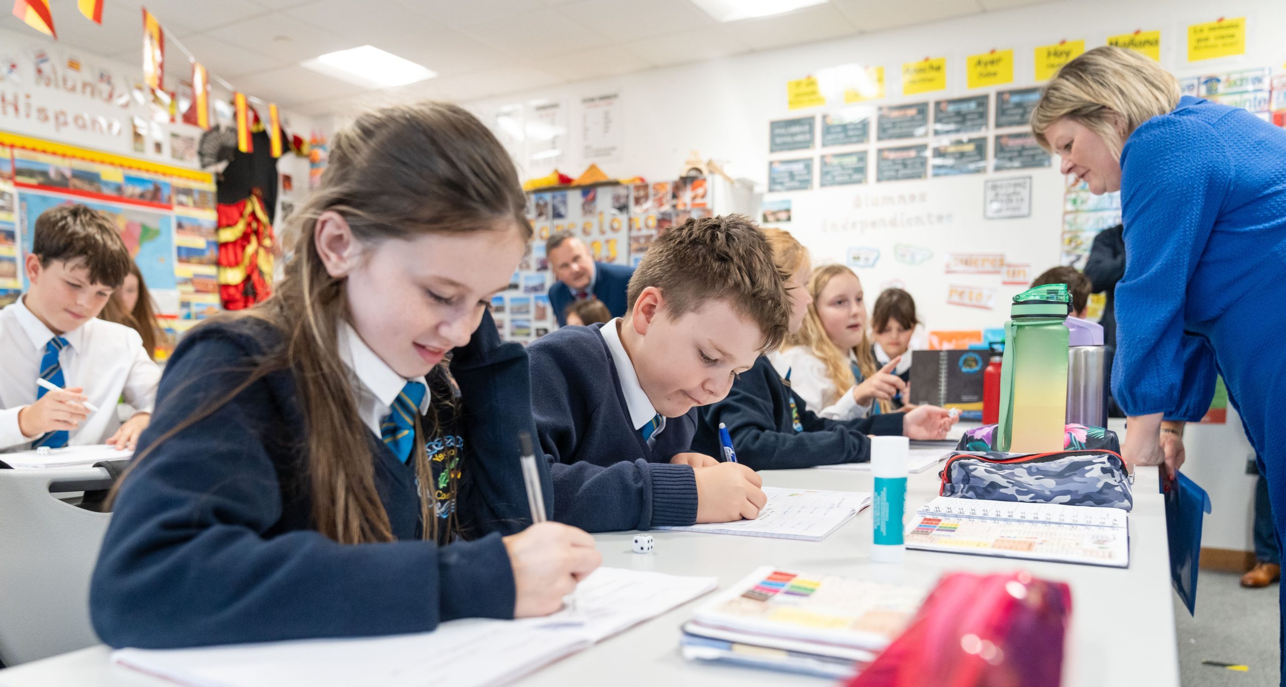 A classroom scene with students in school uniforms working on an assignment. A teacher is leaning over to assist a student. The students are focused, writing in their notebooks, with various school supplies on the desk. The classroom walls are decorated with colorful posters and flags.