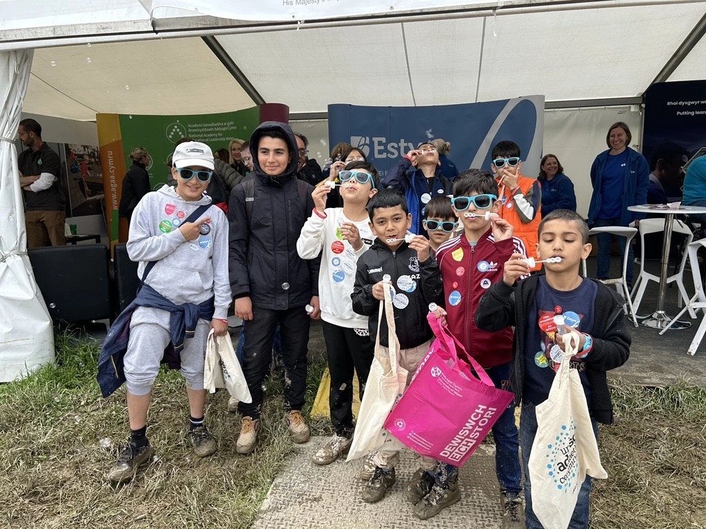 A group of children, wearing sunglasses and holding bags, stand in front of a tent at an outdoor event. They are smiling and display various colourful stickers on their clothes.