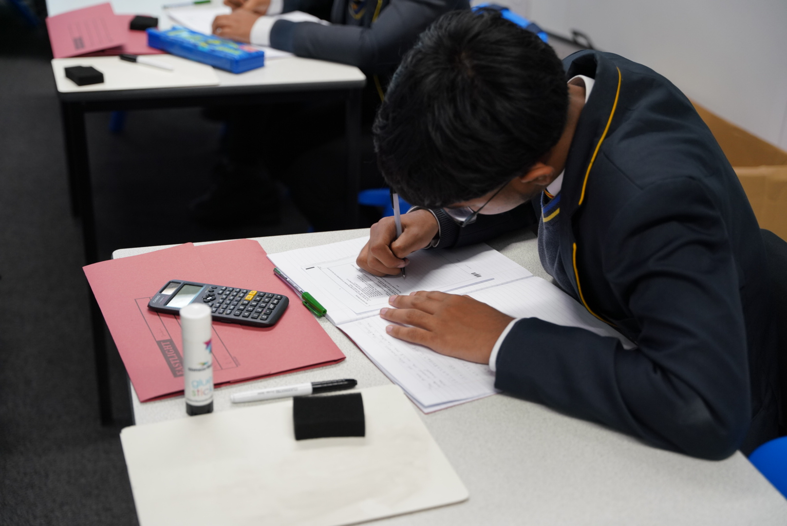 A student in a school uniform working at a desk, focused on writing in a notebook with a calculator, glue stick, and folder nearby