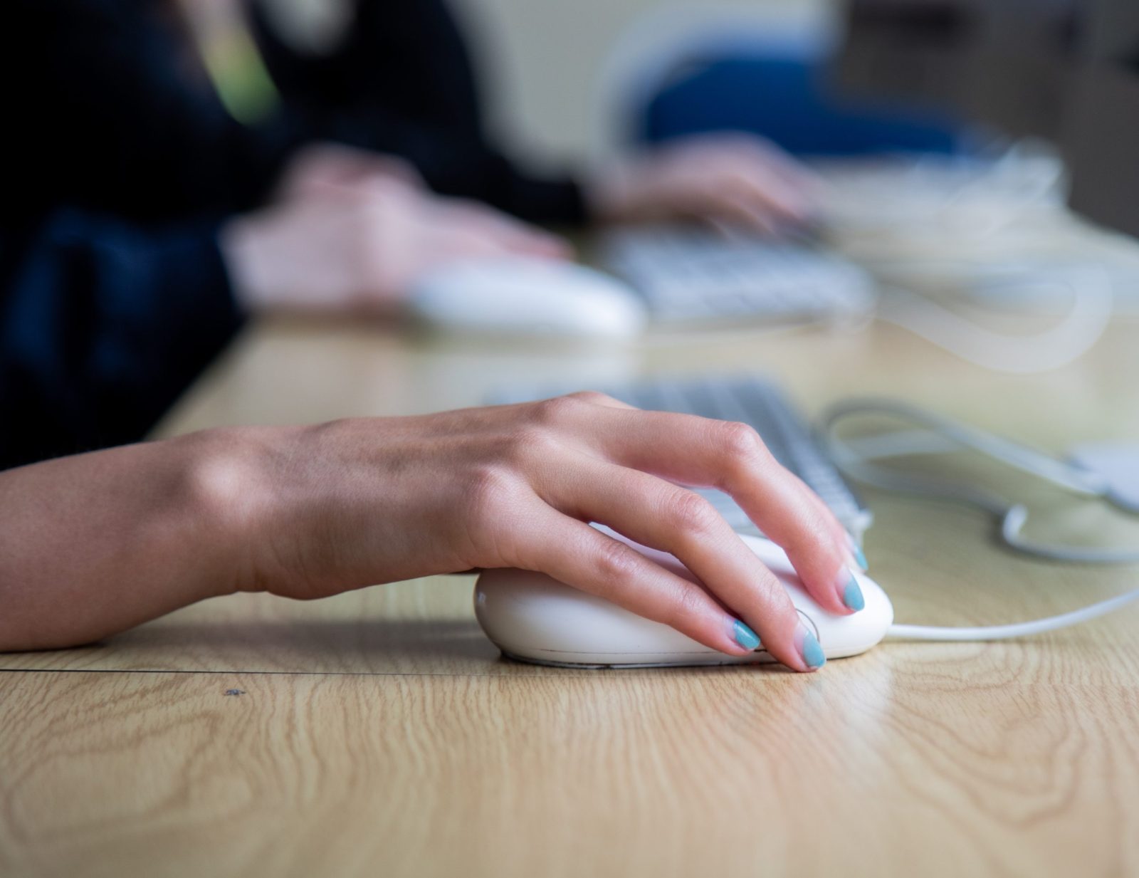 A close-up shot of a hand with blue-painted nails using a computer mouse, with another person in the background working on a computer keyboard.