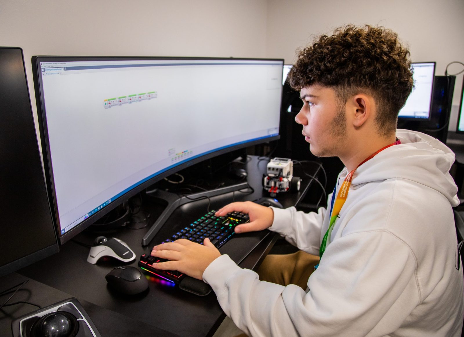 Student working on a computer with a large, curved screen, focused on programming or coding in a classroom setting.