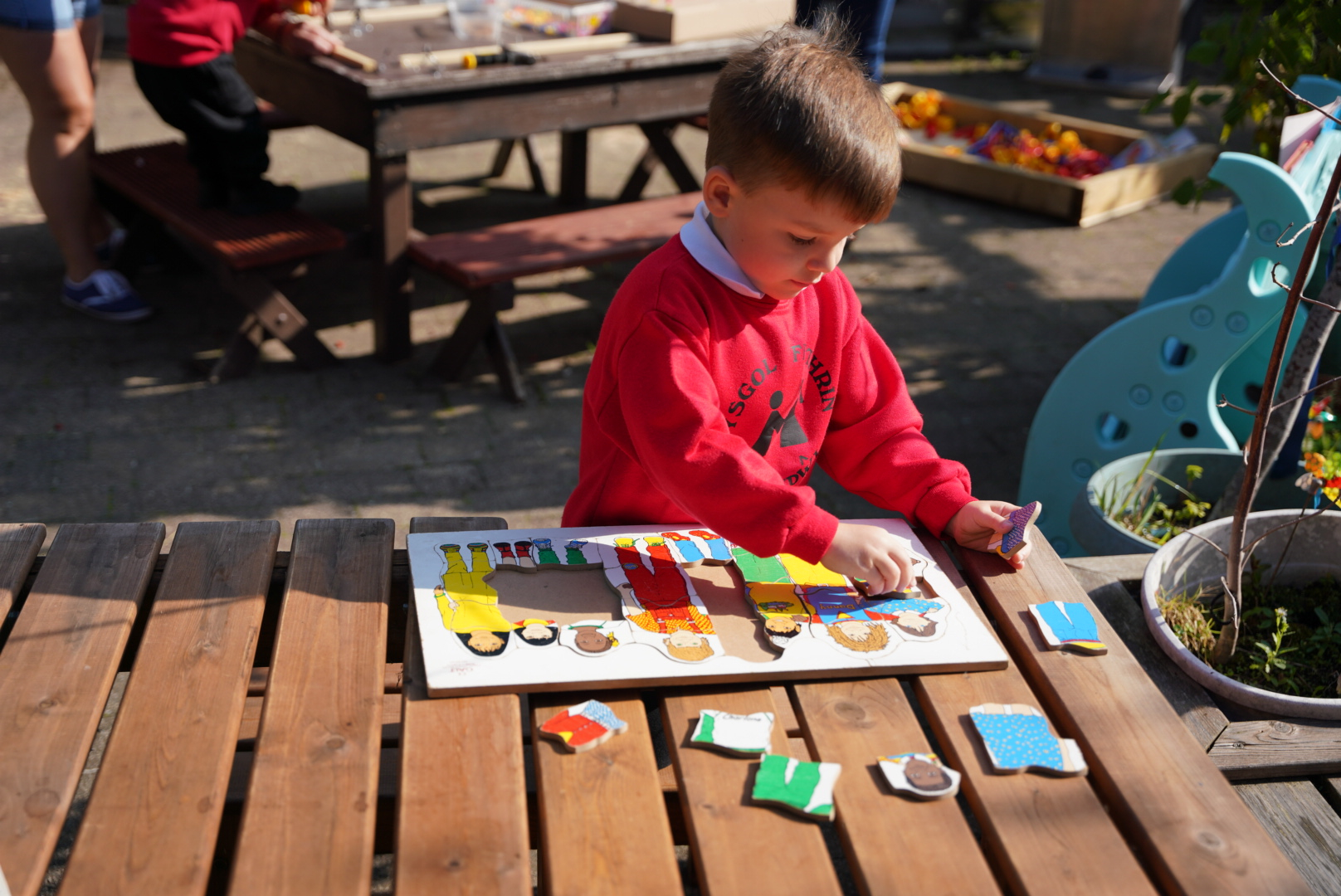 A child in a red school jumper concentrating while placing puzzle pieces at an outdoor table.