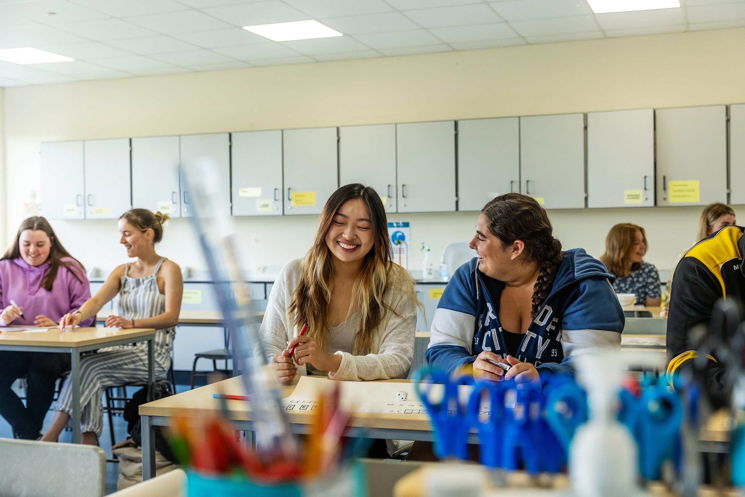 Students are sitting at desks in a classroom, smiling and engaged in conversation. The atmosphere appears to be relaxed and friendly, with classroom supplies visible in the foreground.