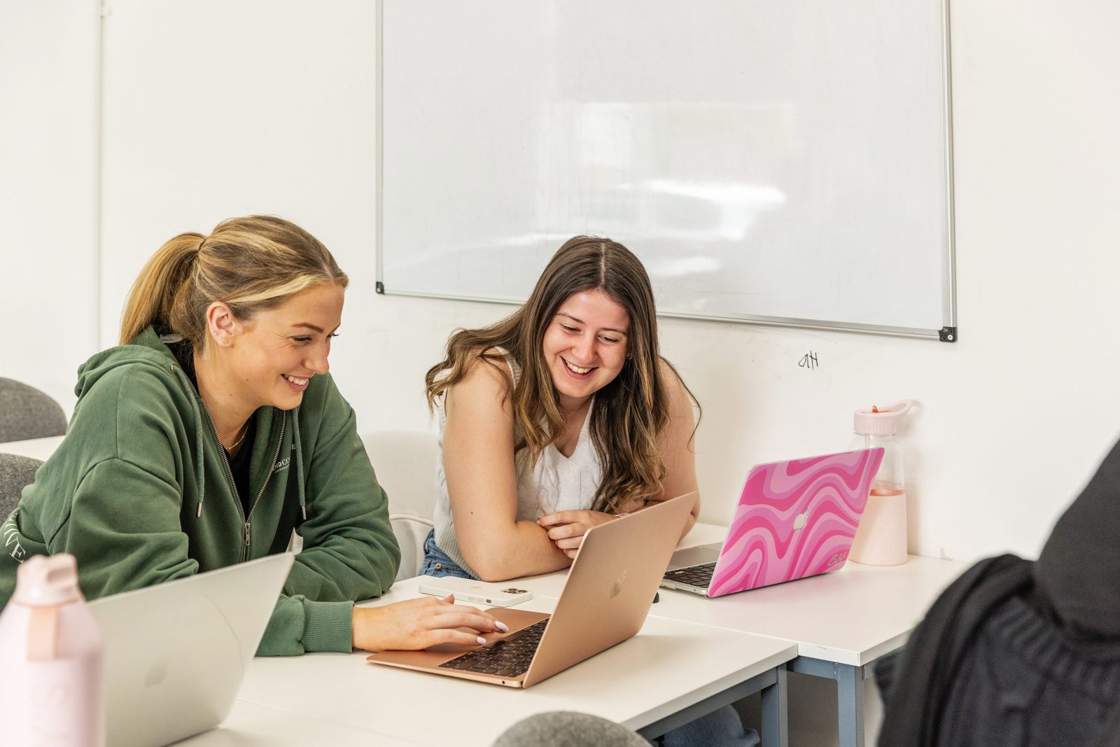 Two female students sitting at a table with laptops, smiling and engaged in conversation in a classroom setting.