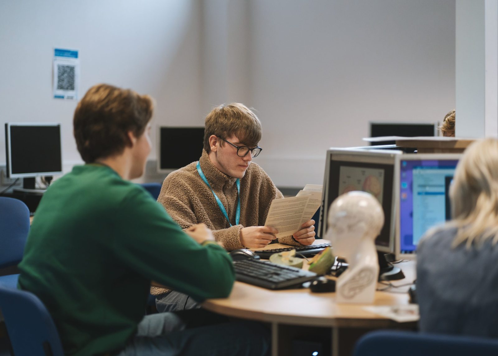 Two students sitting at a round table in a computer lab. One is wearing a green sweater, and the other is reading a document while wearing a light brown fleece.