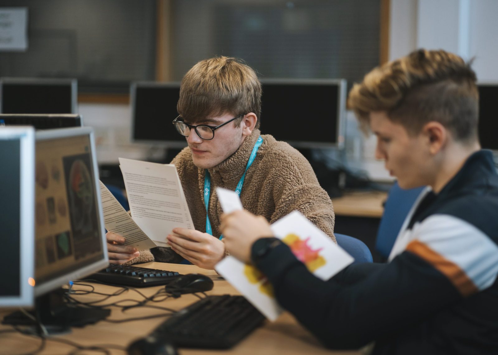 Two students in a computer lab, both reading documents. The student on the left is wearing a light brown fleece, while the student on the right is wearing a navy jacket with white and orange stripes.