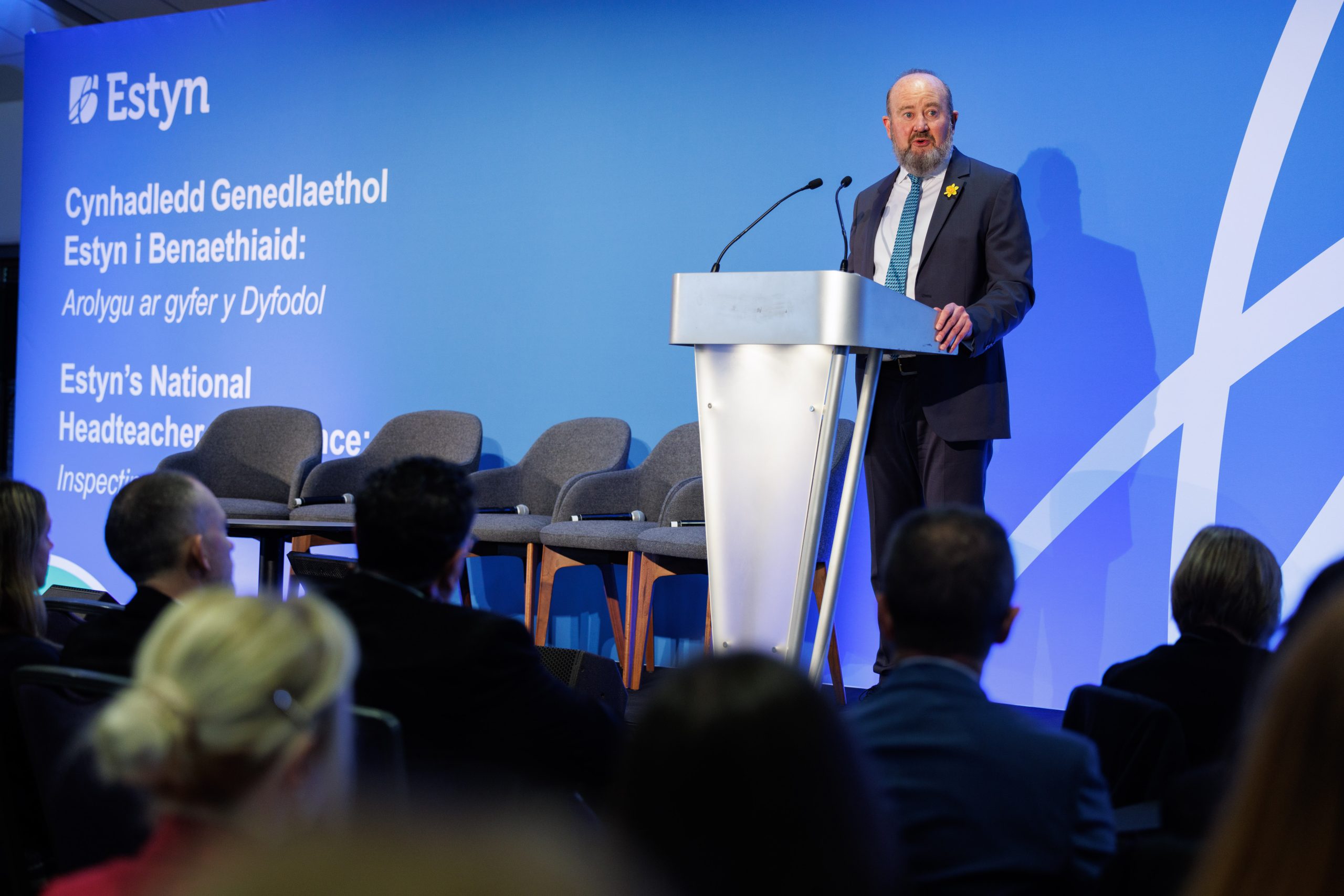 Owen Evans speaks at a podium during Estyn's National Headteacher Conference. The background features the Estyn logo and bilingual text in Welsh and English.