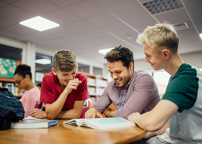 A teacher and two teenage boys laughing while studying in a library.