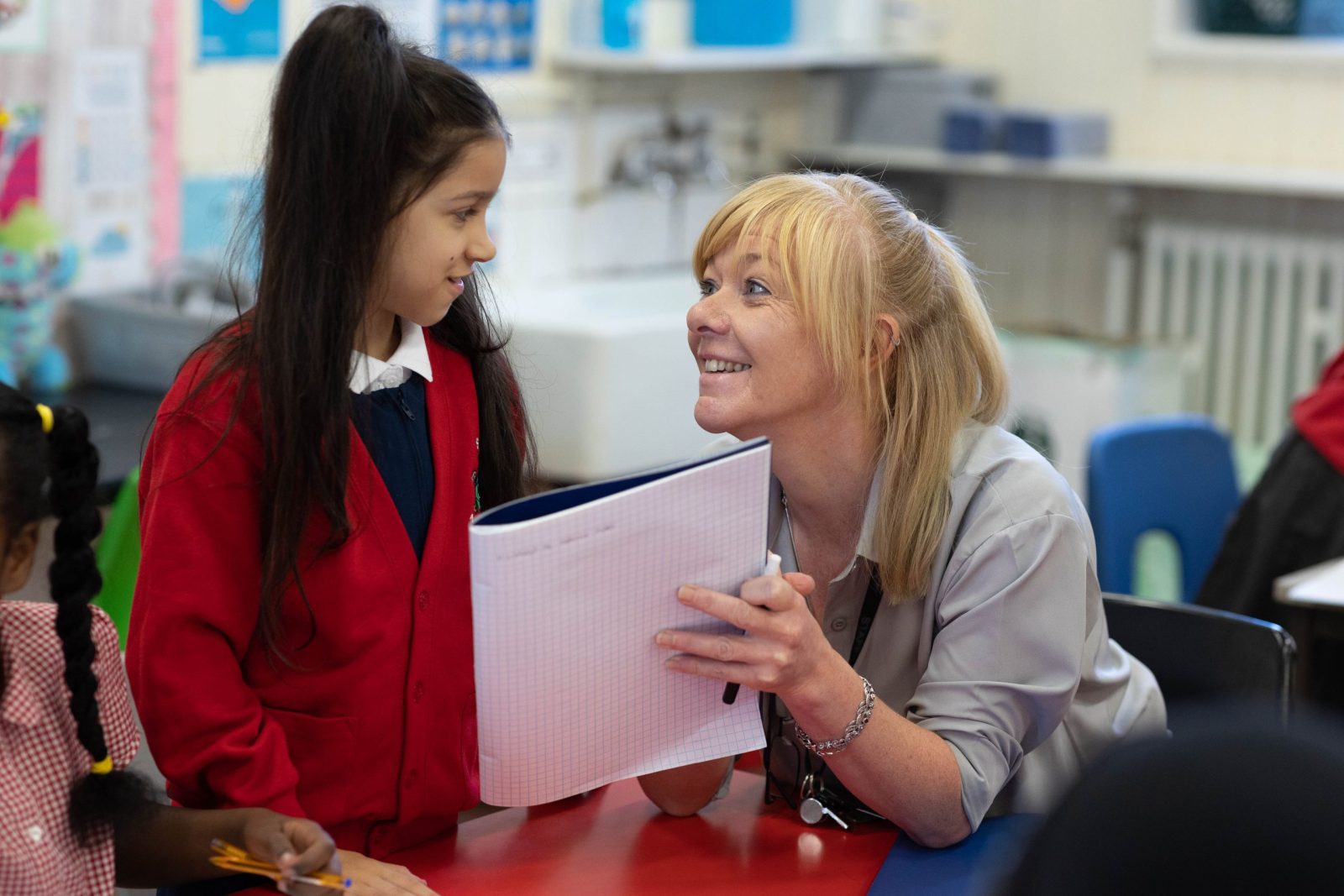 teacher smiling warmly at a young girl in a red cardigan, holding a notebook during a classroom activity.