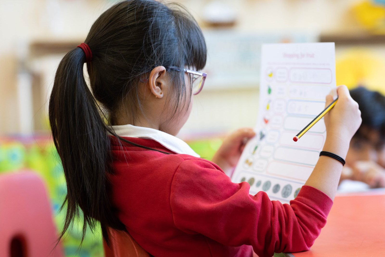 A young girl with long dark hair in a ponytail, wearing a red school uniform, sits in a classroom holding a pencil and looking at a worksheet. The image is taken from behind, focusing on the worksheet and the back of her head.