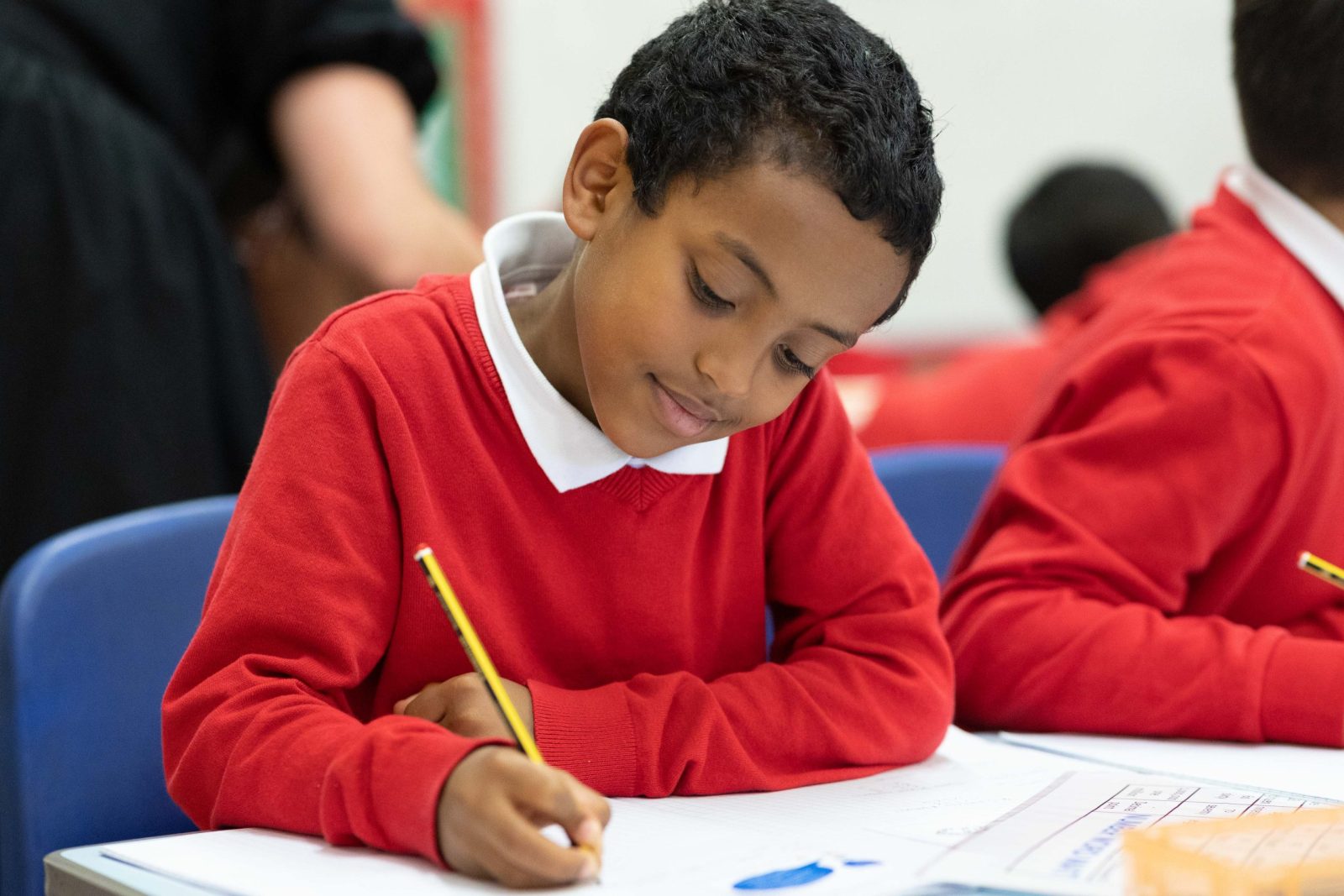 A boy in a red school jumper writes attentively in his notebook during a class activity