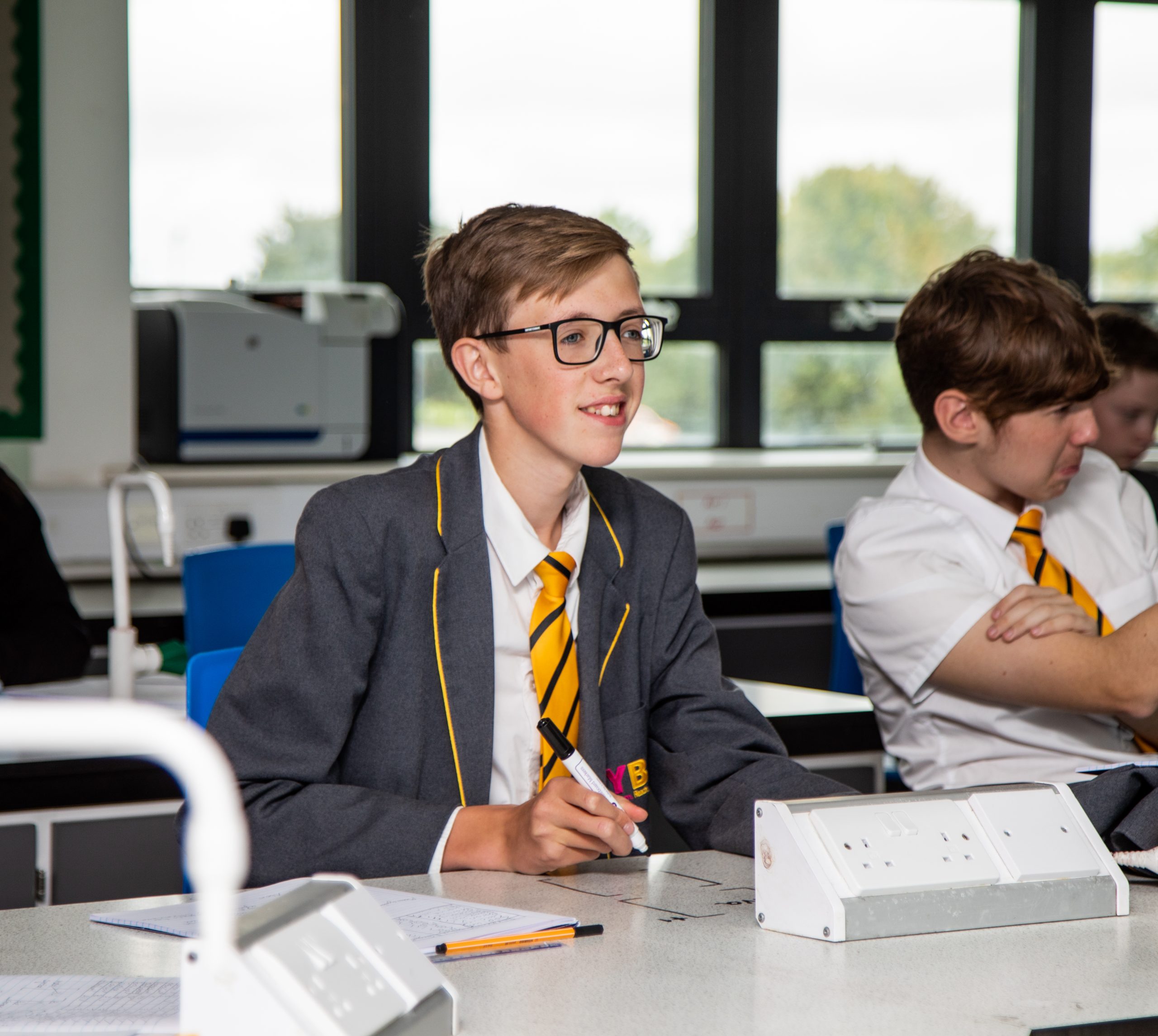 A student in a grey school blazer and yellow tie attentively listening during a class, holding a marker pen, with other students in the background.