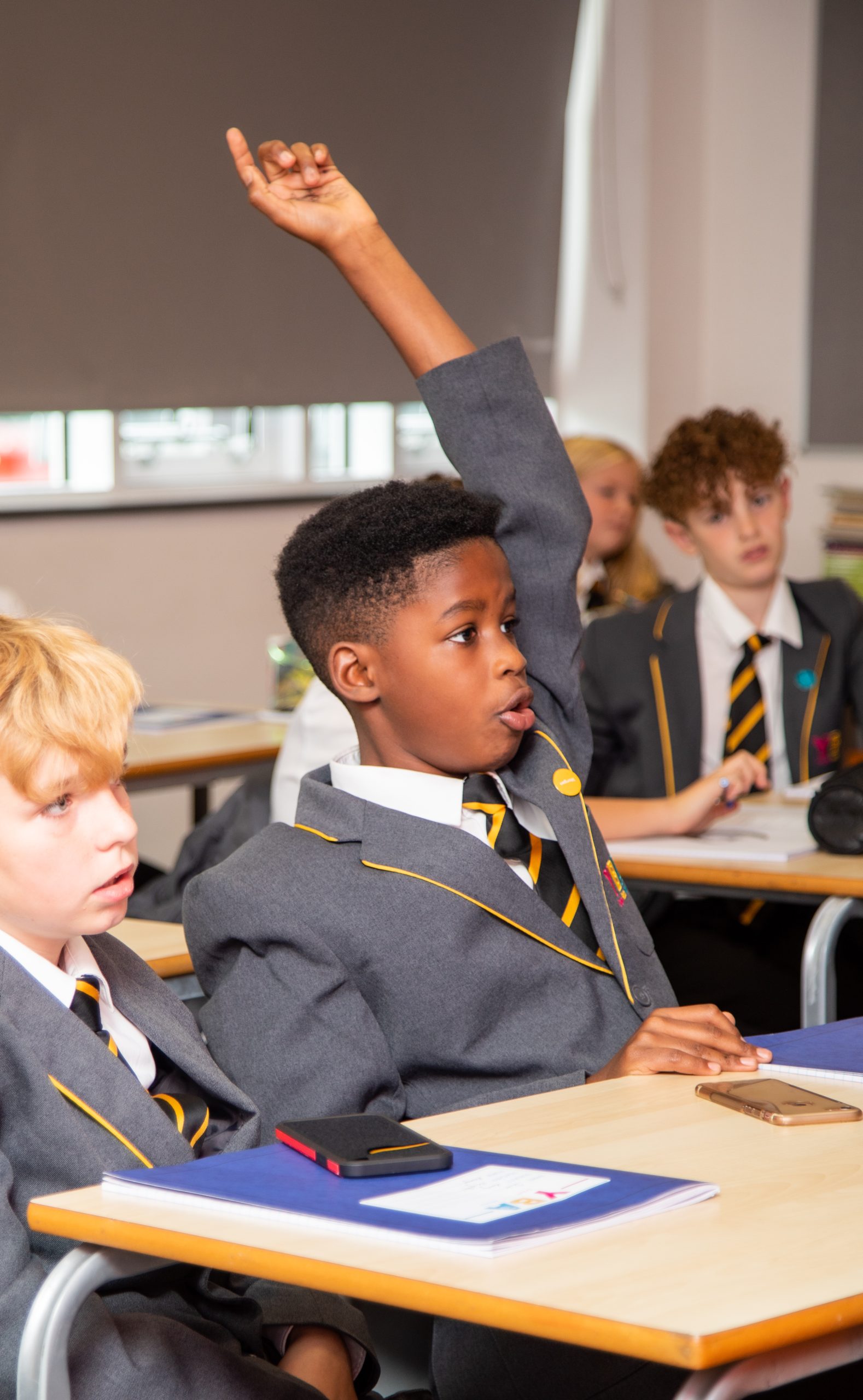 A student in a grey school blazer with a yellow tie raising their hand in class, looking engaged. Other students in similar uniforms are seen in the background.