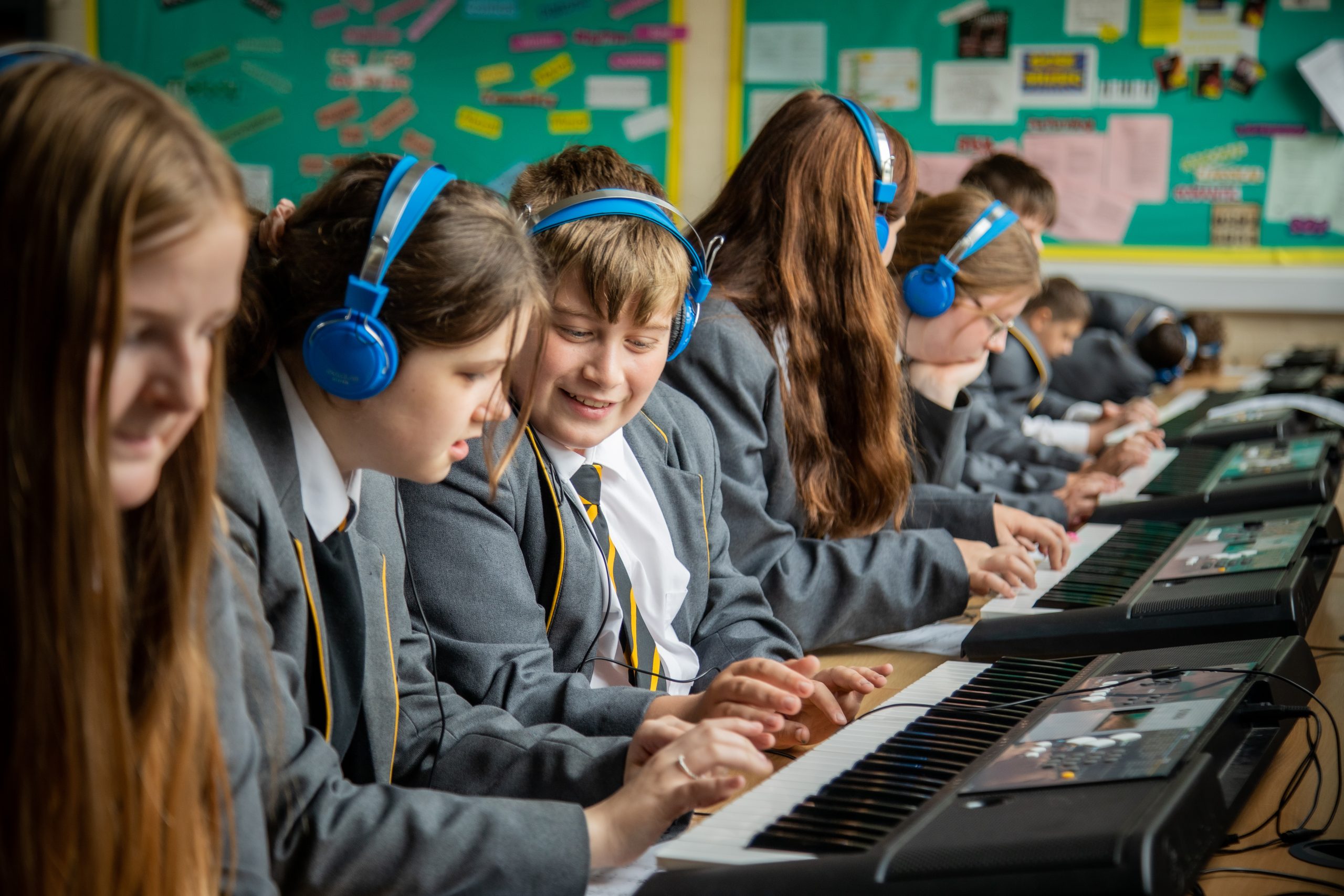 Students in school uniforms are seated in a row, wearing blue headphones and playing electronic keyboards. They are focused on their music lessons, with one student smiling at another as they play. The classroom wall in the background is covered with colorful posters and notes.