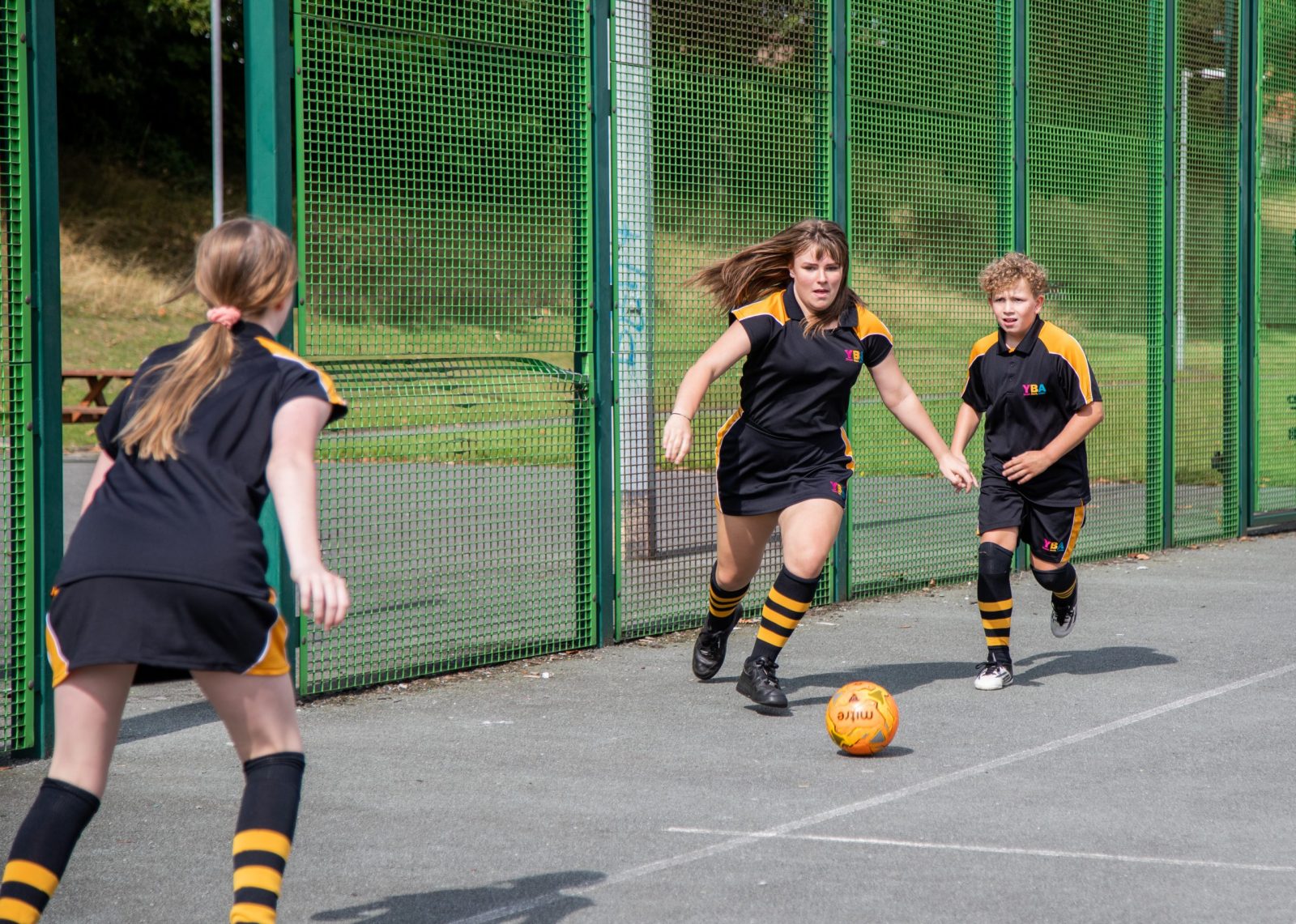 Three students are playing football on an outdoor pitch, wearing black and yellow uniforms.