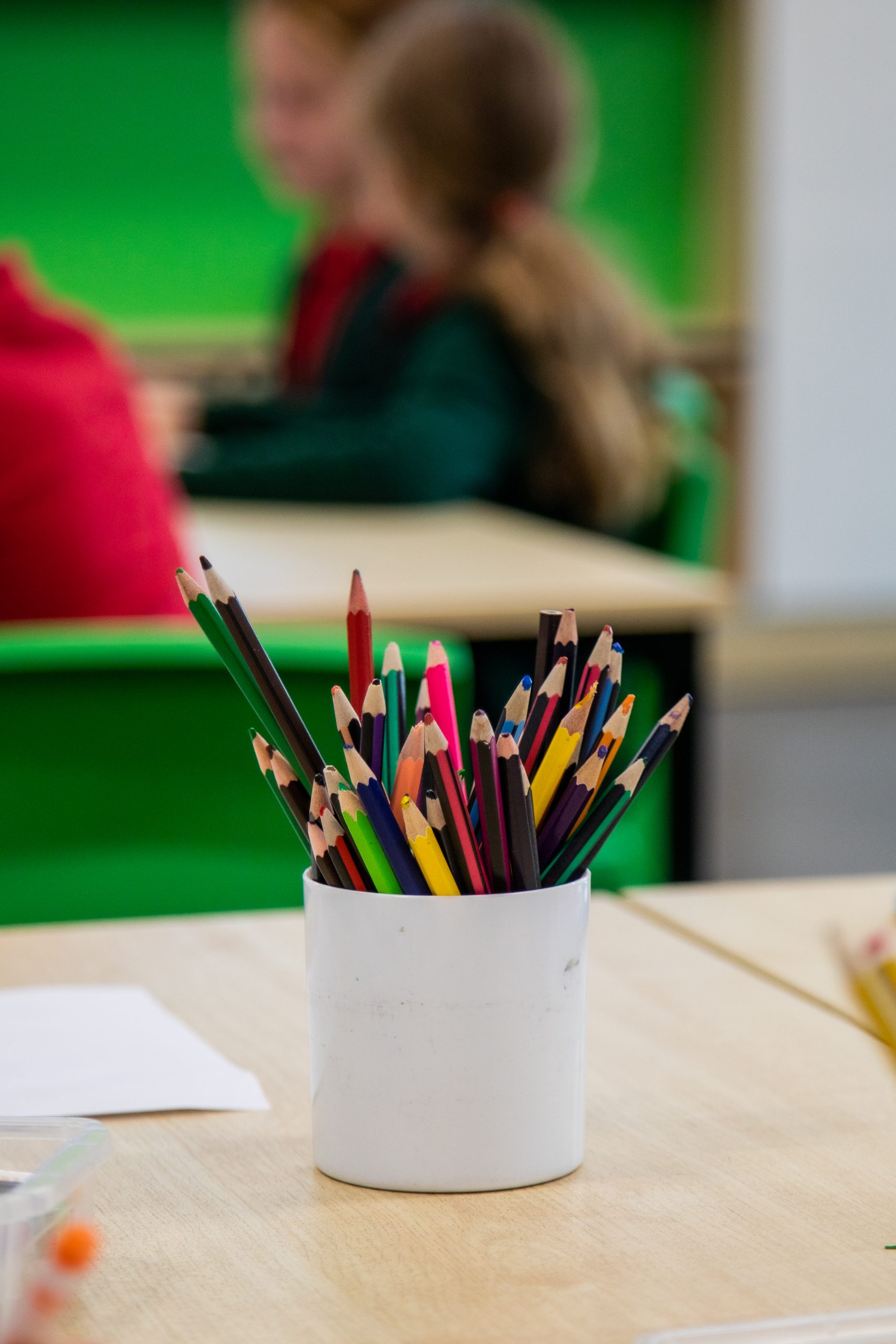 A container filled with colorful pencils on a table, with blurred children in the background.