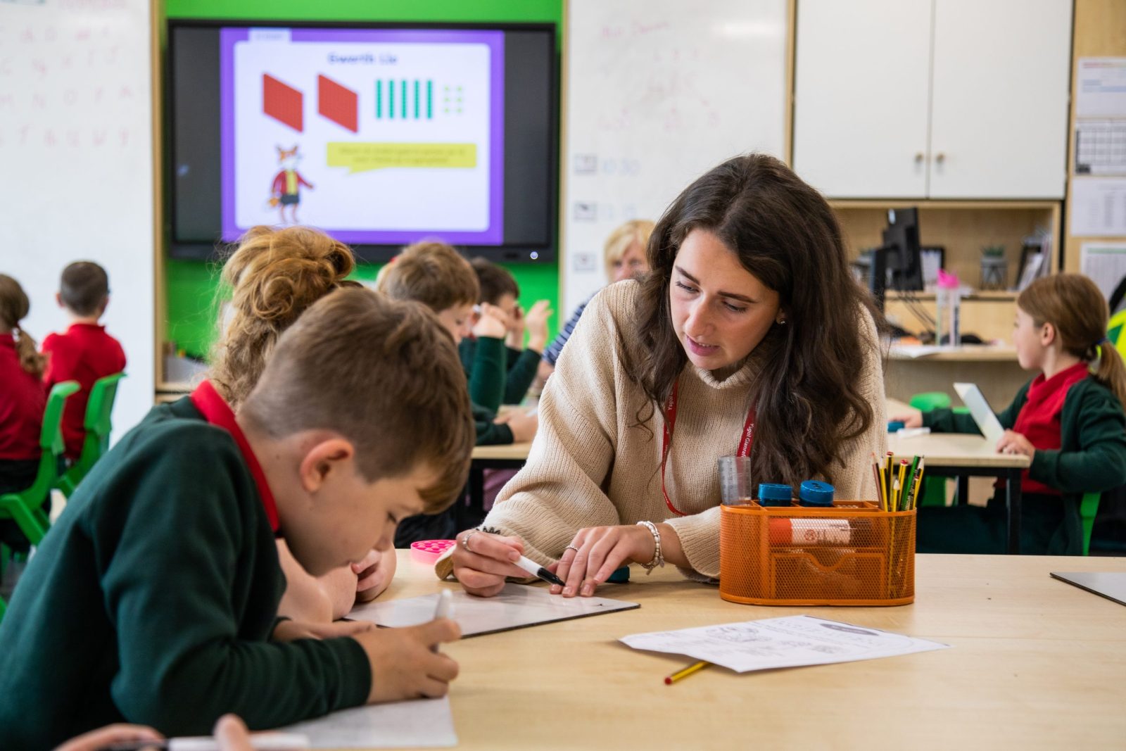 A teacher helping a young student with their work in a lively classroom setting