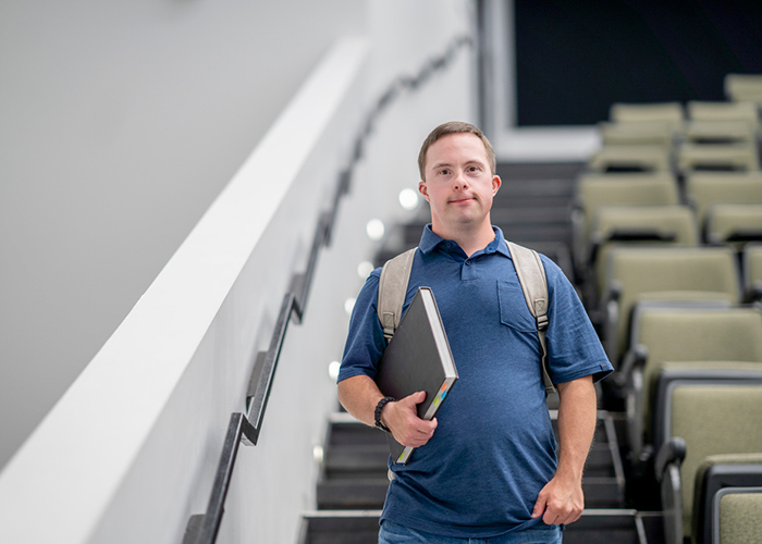 A young man with Down syndrome holding a notebook, standing on stairs in an educational setting.