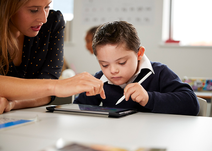 A teacher helps a young boy with Down syndrome use a tablet in a classroom."