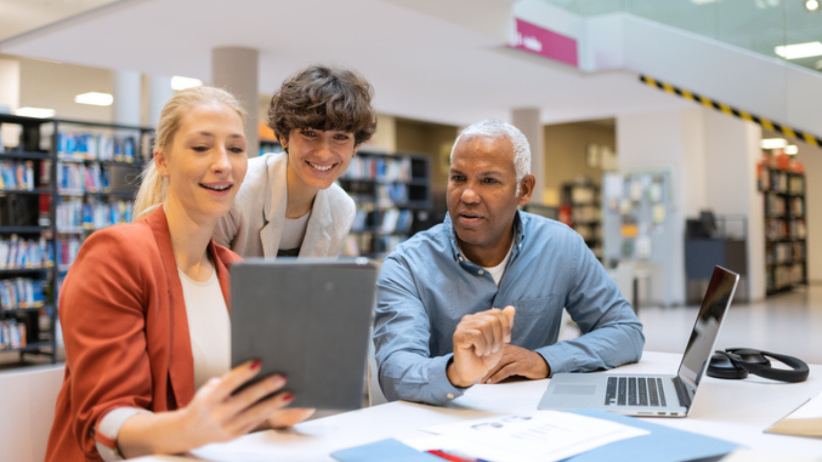 Three people gathered around a laptop in a modern library setting.