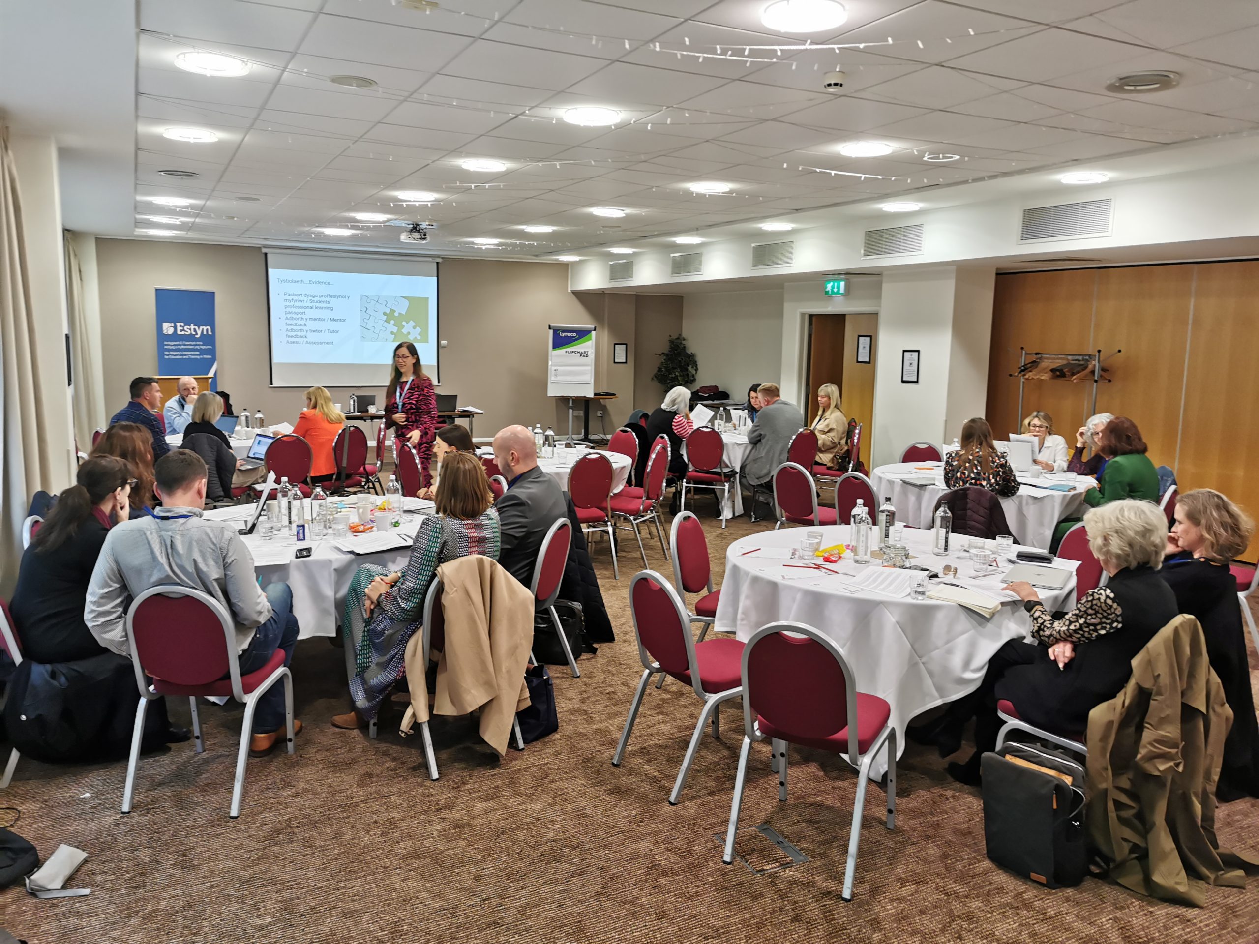Group of attendees seated at round tables in a conference room, listening to a presenter at the front near a screen displaying a PowerPoint presentation.
