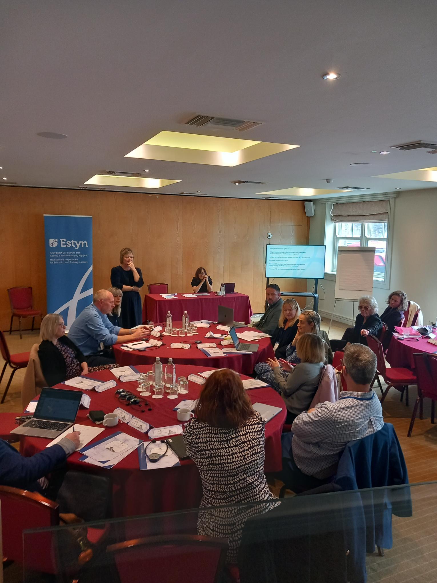 Group of individuals participating in a workshop inside a conference room with Estyn banners displayed.