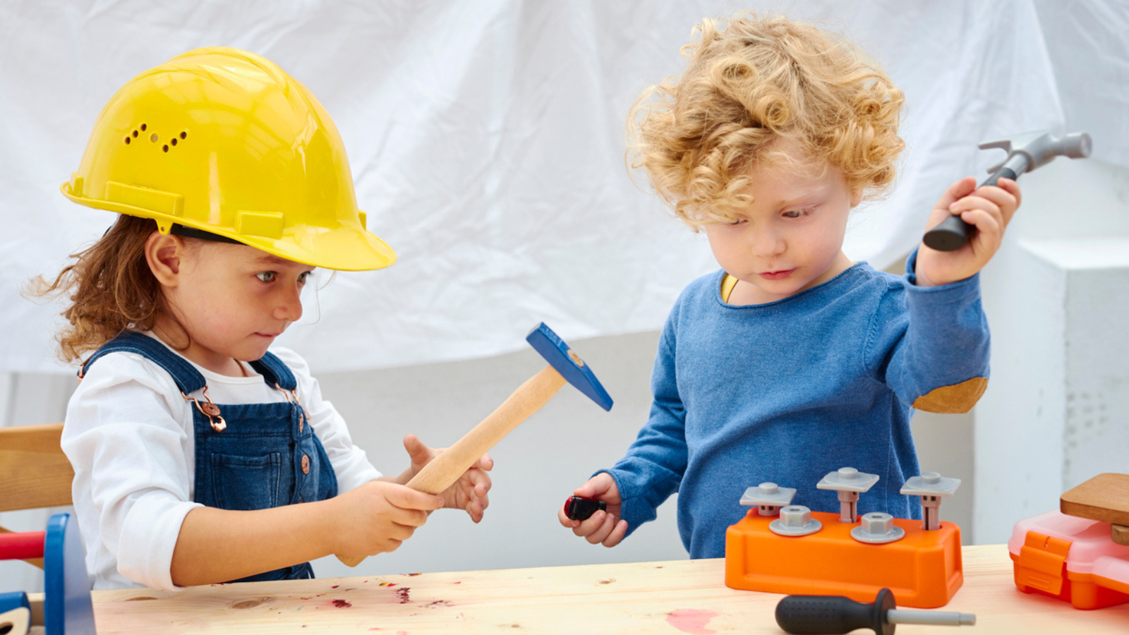 Two children in a workshop setting, with one wearing a yellow hard hat and using a hammer, and the other holding a toy screwdriver. They are surrounded by various tools and a toy workbench.