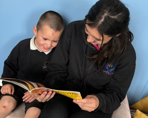 Young child and young person sitting and reading a book together, smiling and engaged in the activity.
