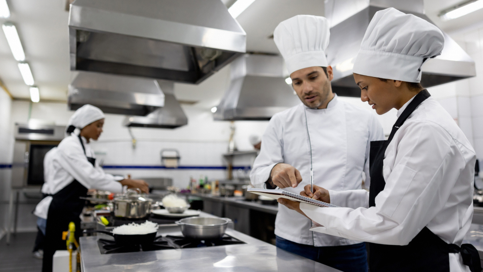 Three chefs in a commercial kitchen, one reviewing a recipe while two others prepare meals.