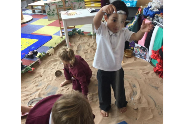 Child playing with a sieve in a sandbox in a classroom setting, with another child nearby.