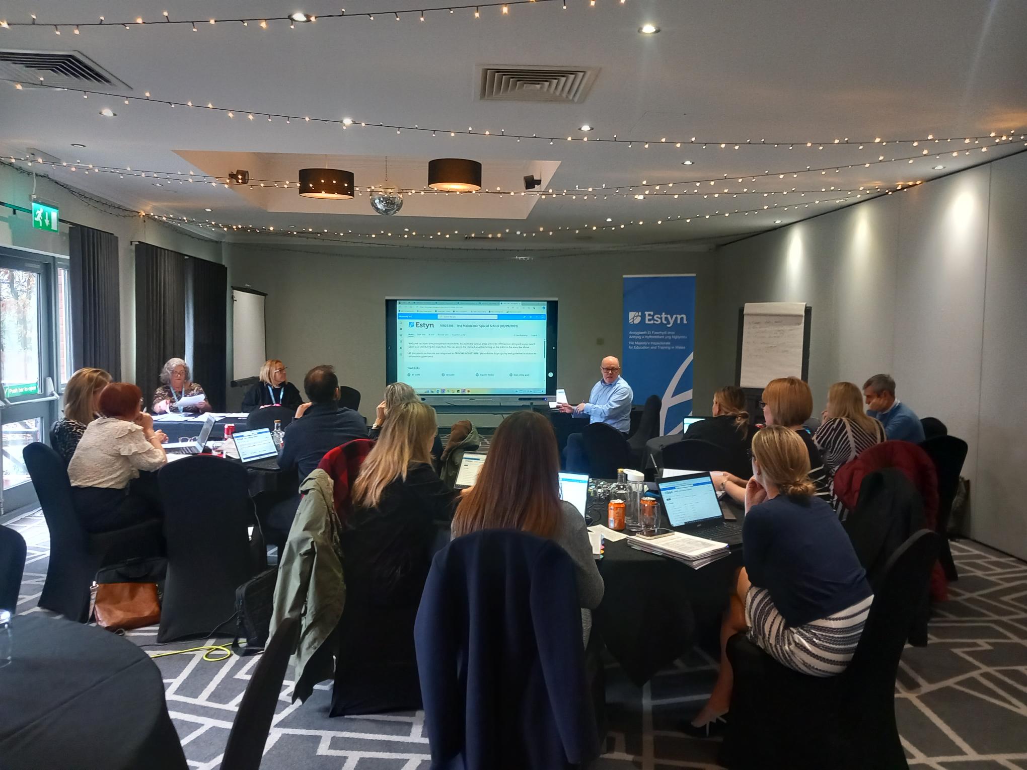 A business workshop with attendees seated around tables, focusing on a presentation that features a digital screen with graphics and text. The setting is indoors, and there is a banner on the right with the name "Estyn" visible.