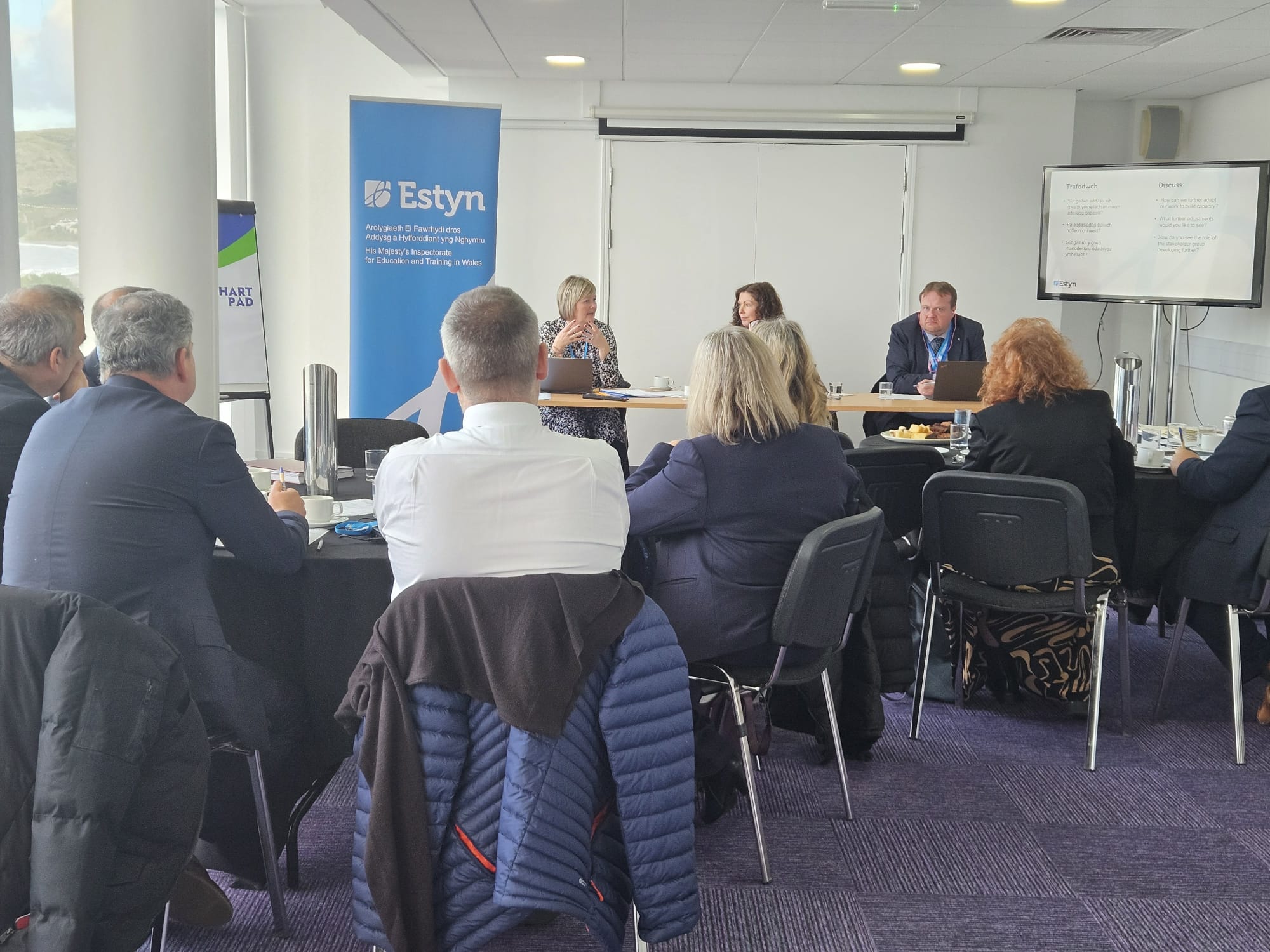 Attendees at a professional meeting with an Estyn banner in the background.