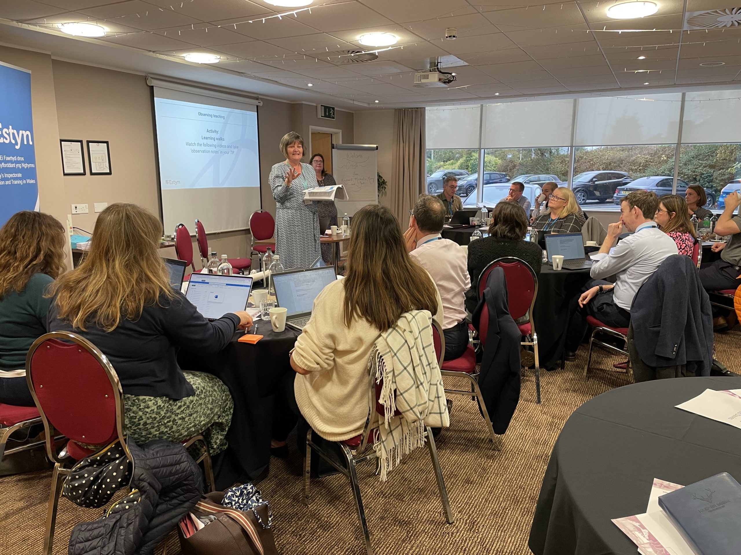 A person presenting at a conference in a well-lit room filled with attendees seated at tables with laptops. Banners with the names "Estyn" are visible in the background.