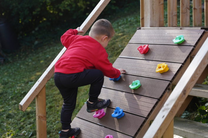 A child in a red jumper is climbing a wooden climbing wall in a backyard.