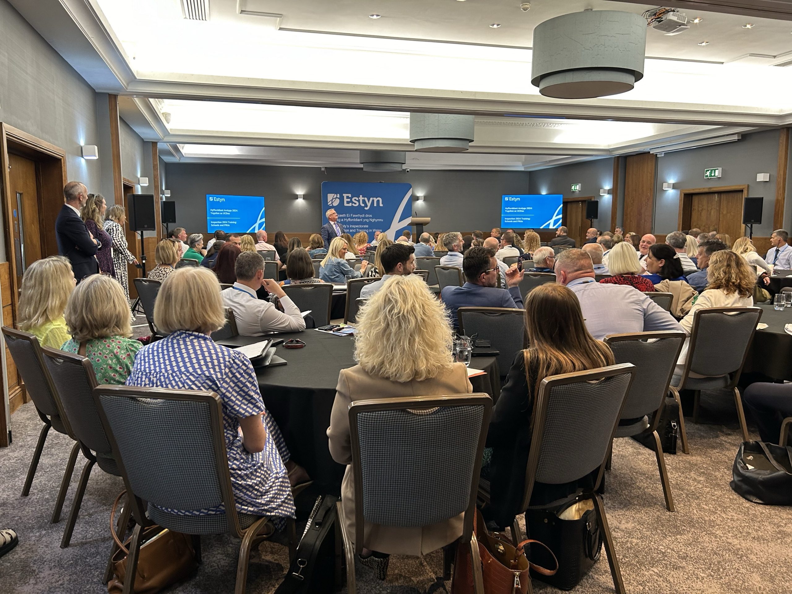 Conference attendees at an Estyn event seated facing two large presentation screens.