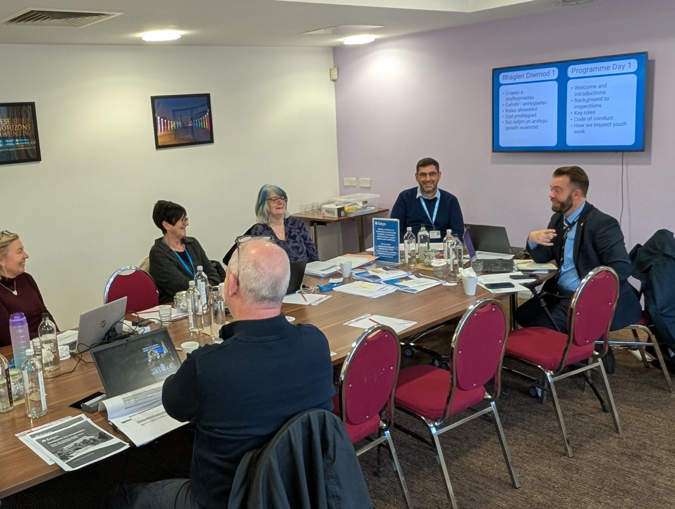 A group of individuals is engaged in a discussion around a table in a meeting room with presentations displayed on screens.