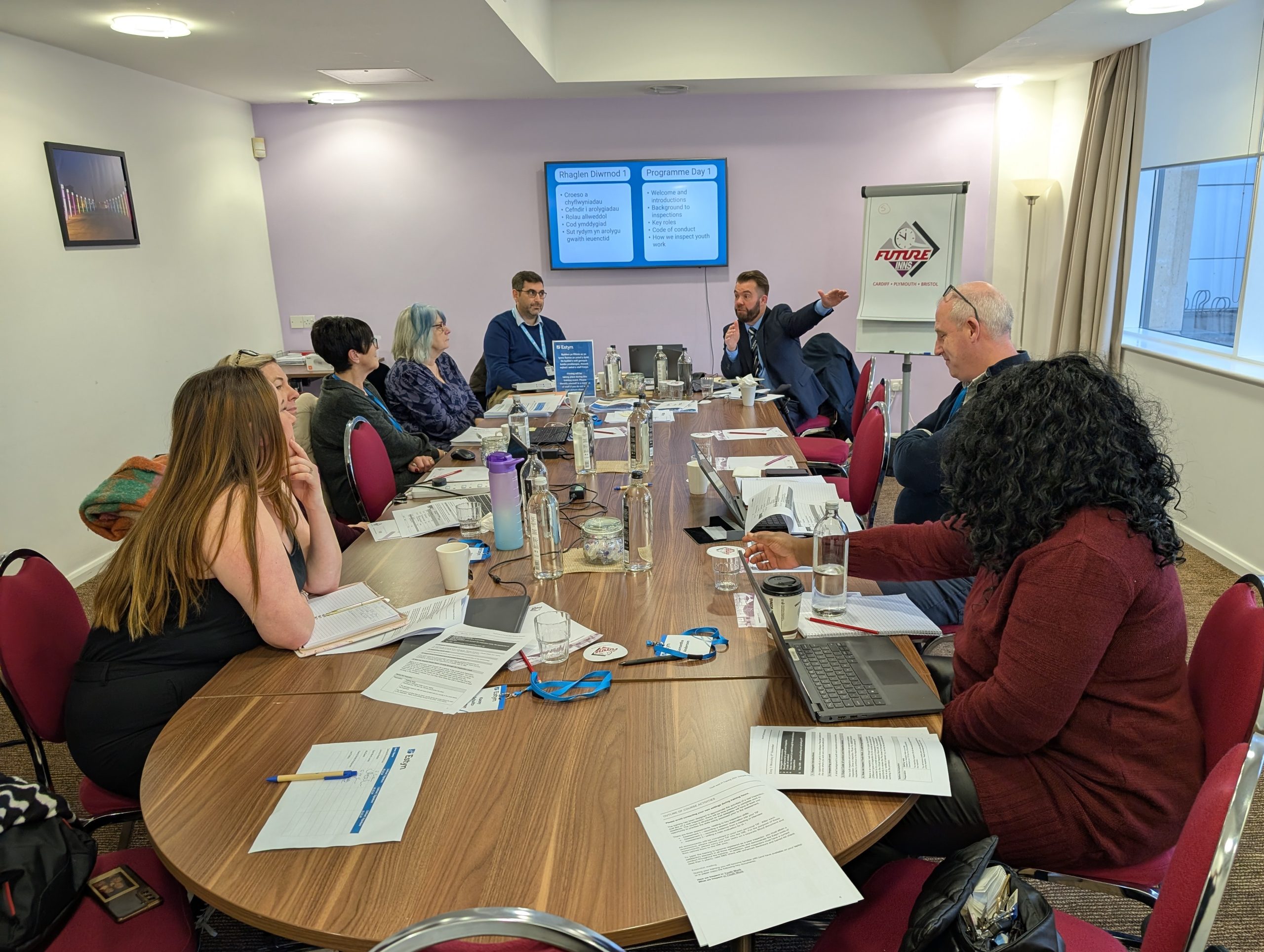 Several individuals in a business meeting around a table, with documents and laptops visible, focused on a presentation about 'Project Efficiency' displayed on a screen at the front of the room.