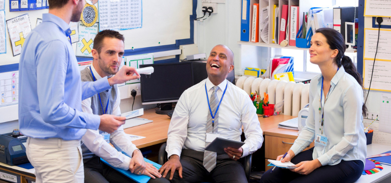 A group of four educators, wearing ID badges, are engaging in a lively discussion in a classroom adorned with educational posters and student work.