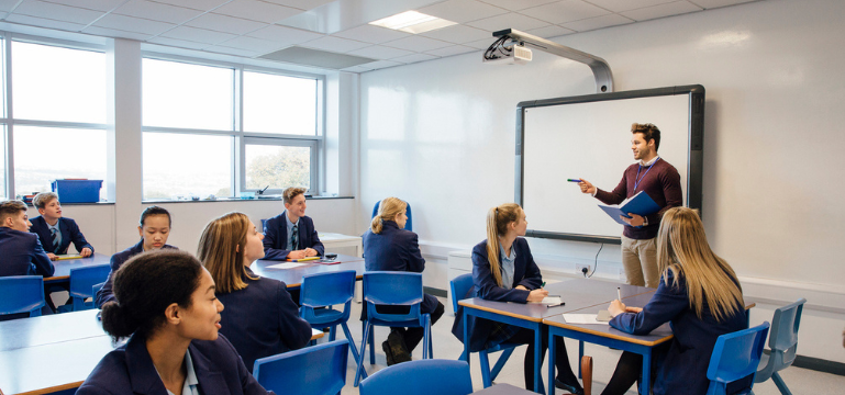 A classroom scene with students in uniform seated at desks and a teacher at the front, discussing a topic near a whiteboard.