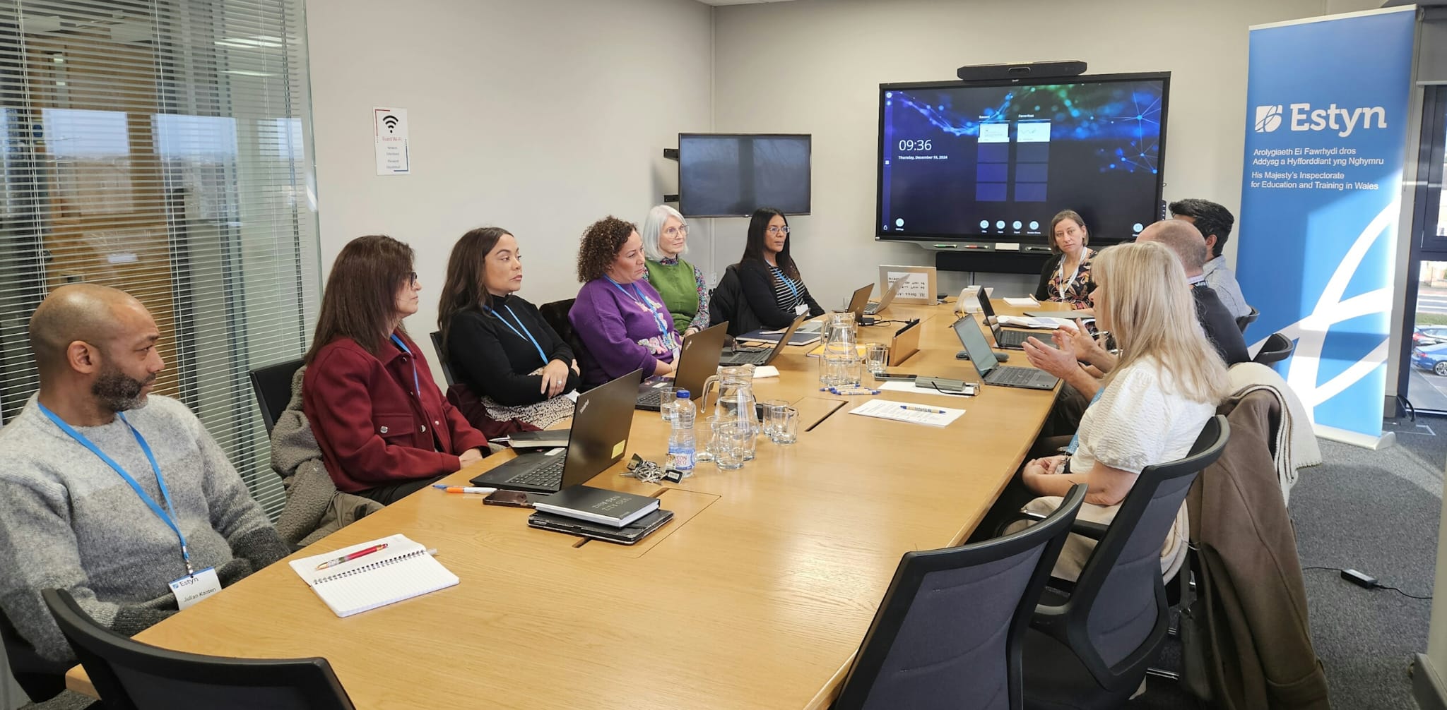 Group of professionals sitting around a conference table in a meeting room with the logo of Estyn on a screen in the background.