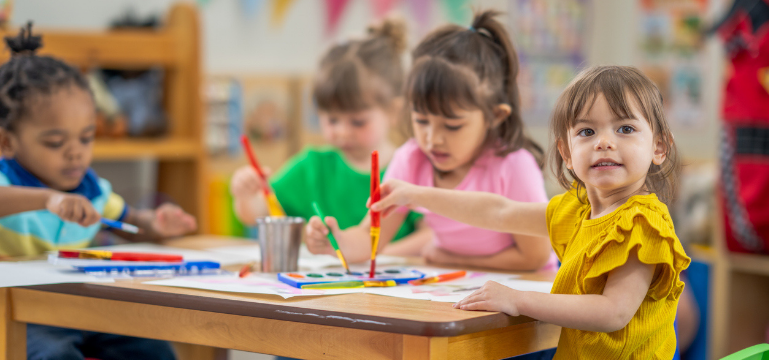 Children engaged in a painting activity at a classroom table, with one child in a yellow shirt looking at the camera, holding a paintbrush.