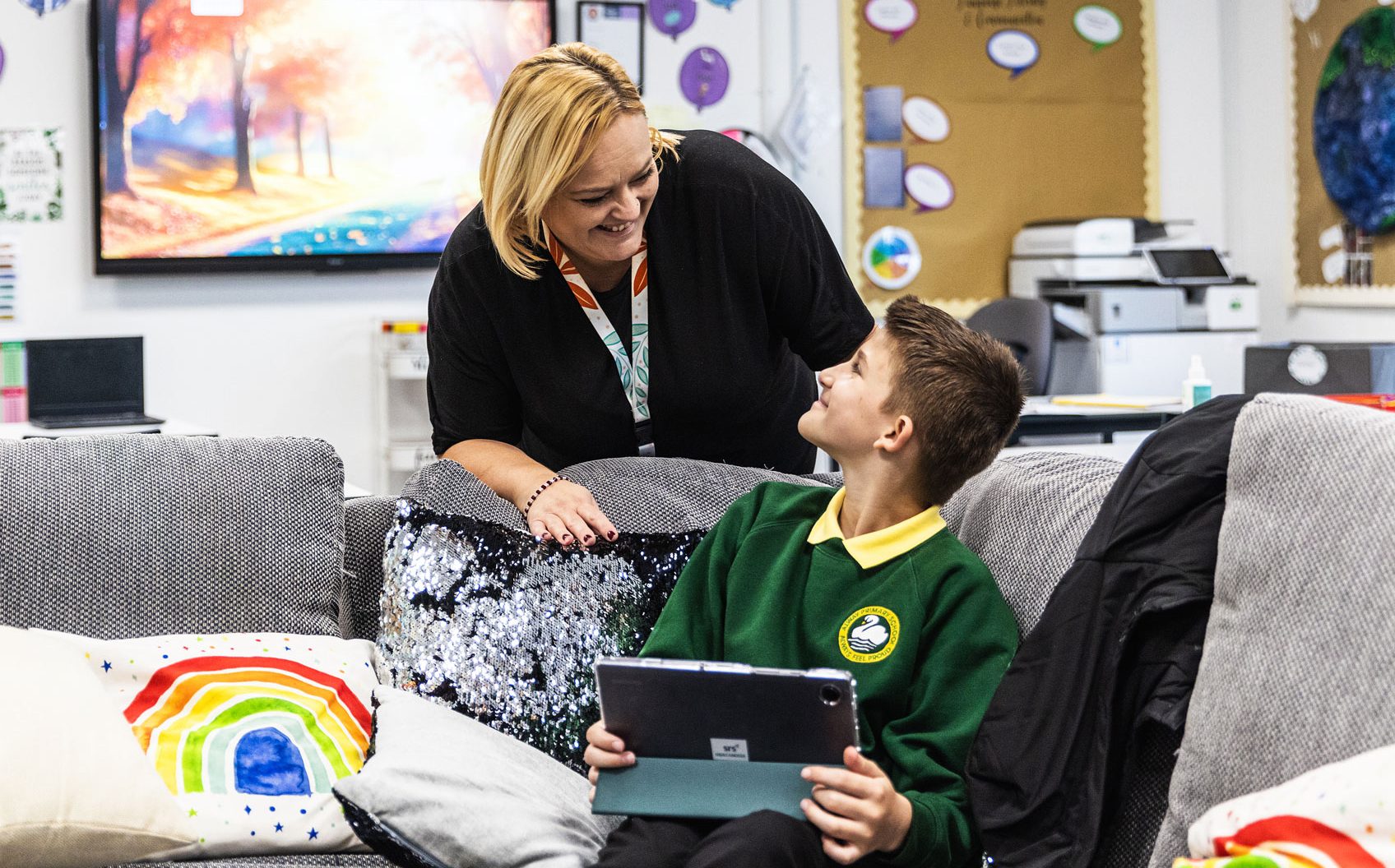 A teacher smiling and leaning towards a student holding a tablet in a cozy classroom with colorful decorations and a relaxing environment.